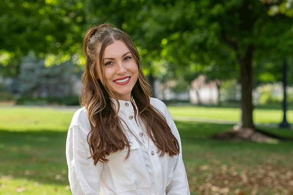 A young female with long brown hair smiling at the camera and standing in the quad in summer with green grass and trees visible.